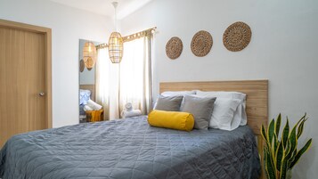Una cama bien hecha con colcha gris, cabecera de madera y almohada amarilla. Hay decoraciones de pared tejidas y una planta en maceta a un lado.
