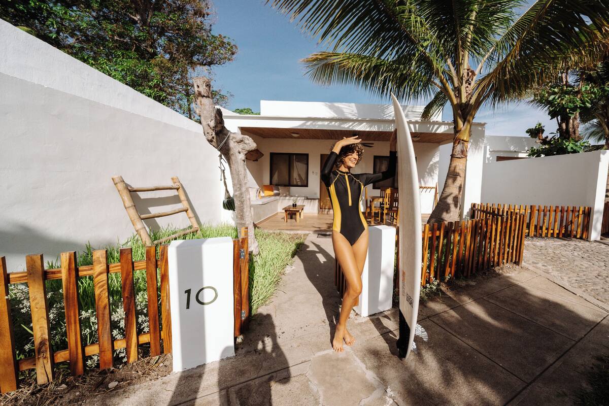 A person in a swimsuit standing next to a surfboard, with a wooden fence and a palm tree in the background.