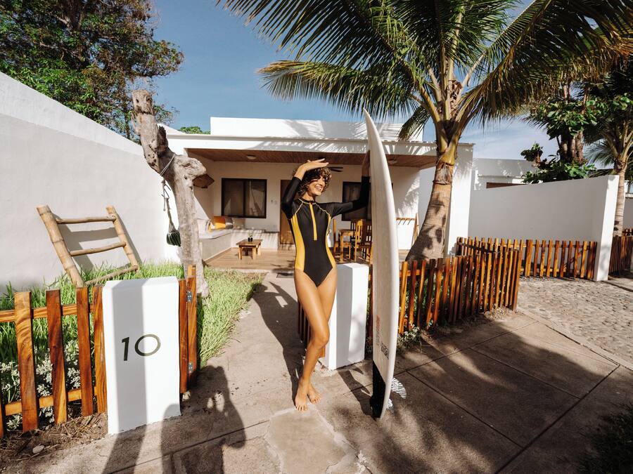 A person in a swimsuit standing next to a surfboard, with a wooden fence and a palm tree in the background.