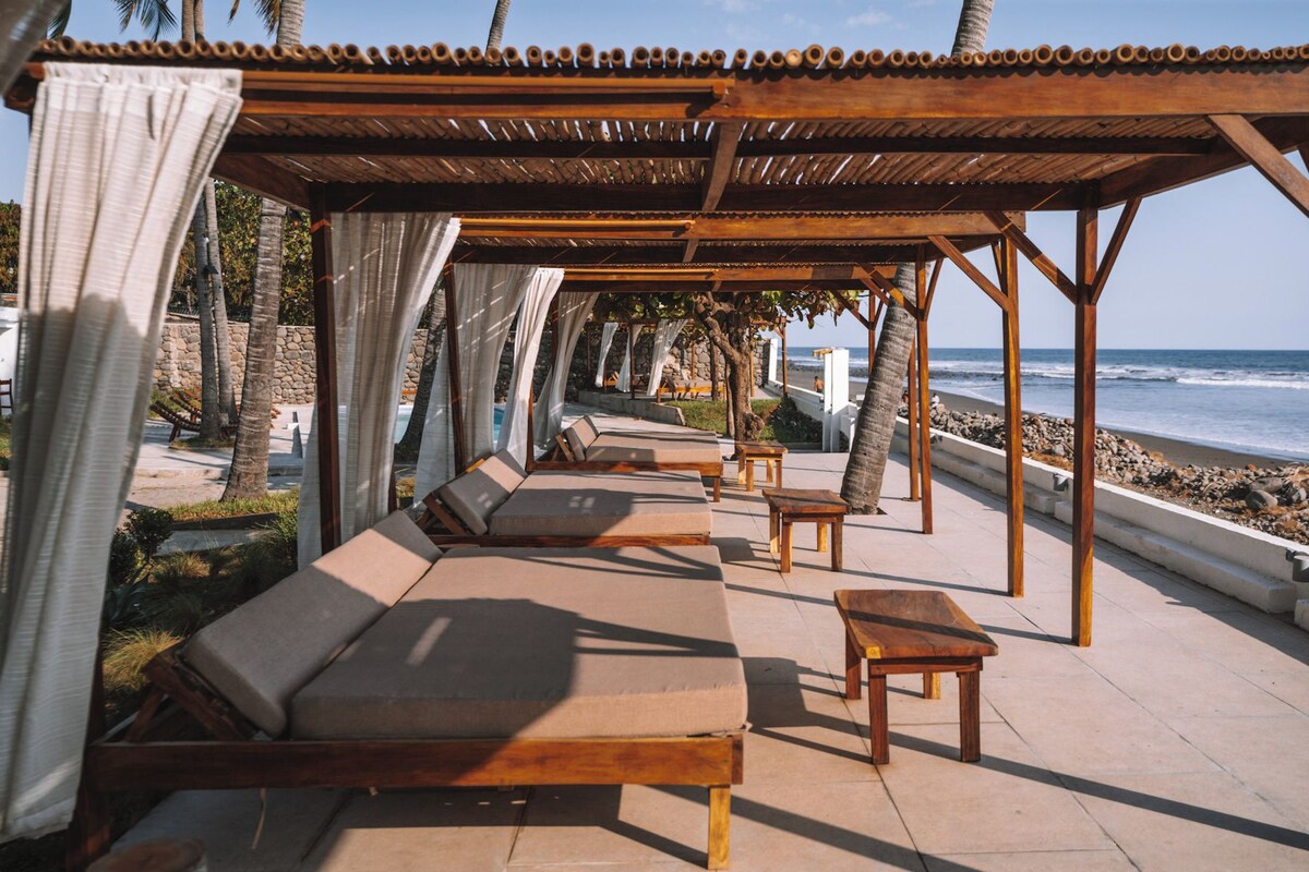 A beachside lounge with wooden loungers and tables, a pergola with a tiled roof, and a view of the ocean.