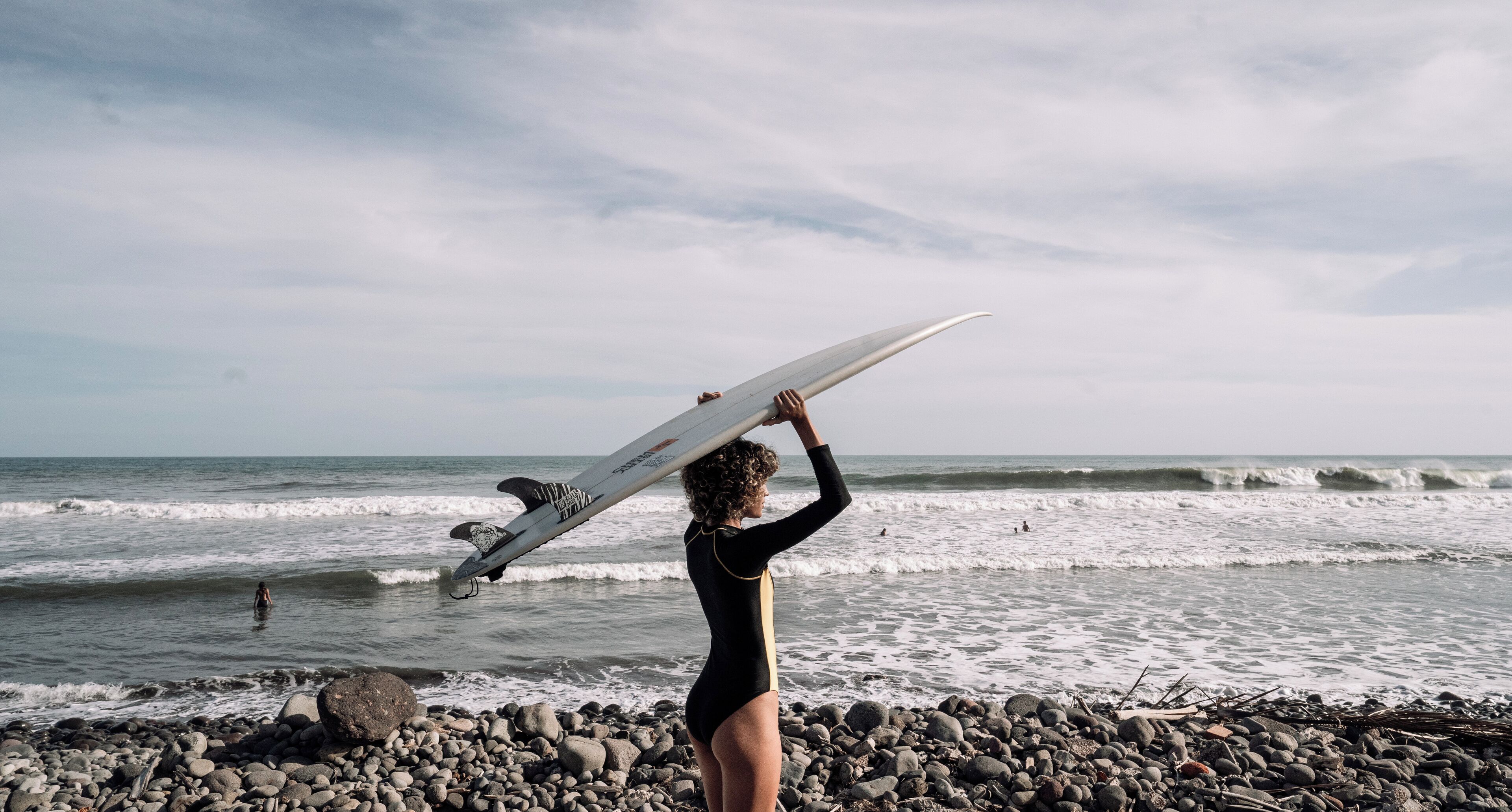 Eine Person hält ein Surfbrett an einem felsigen Strand, im Hintergrund sind Wellen zu sehen.