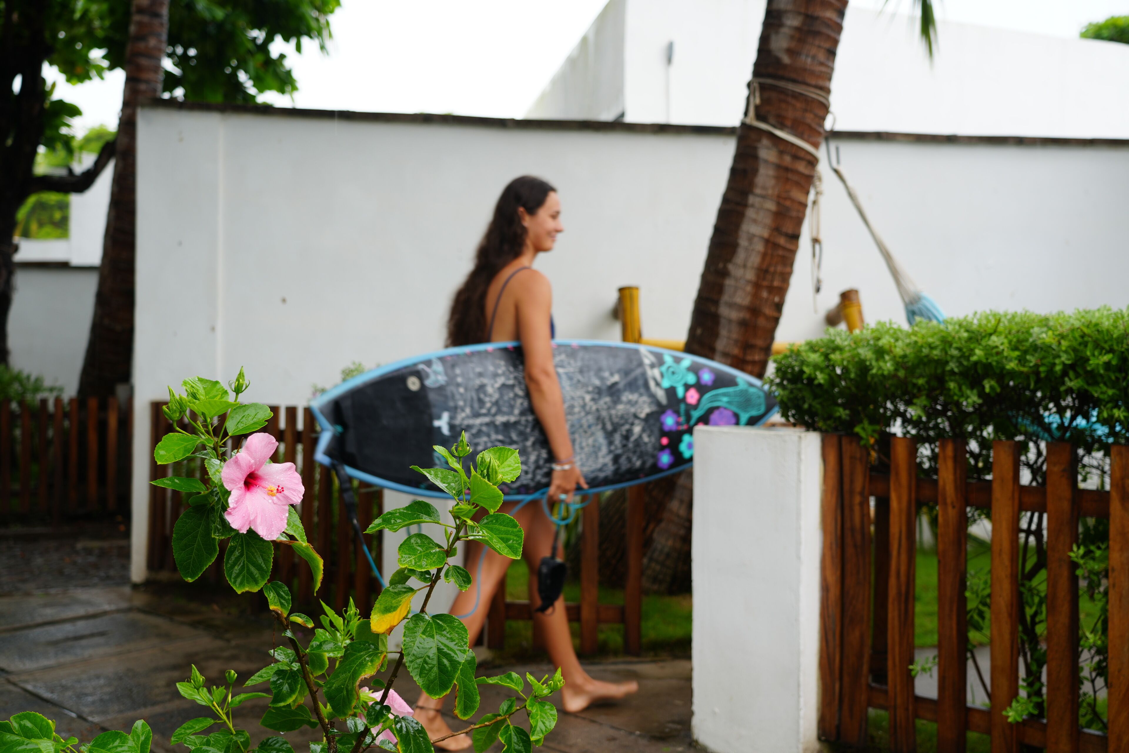 Une personne marchant avec une planche de surf, une clôture en bois et un mur blanc.