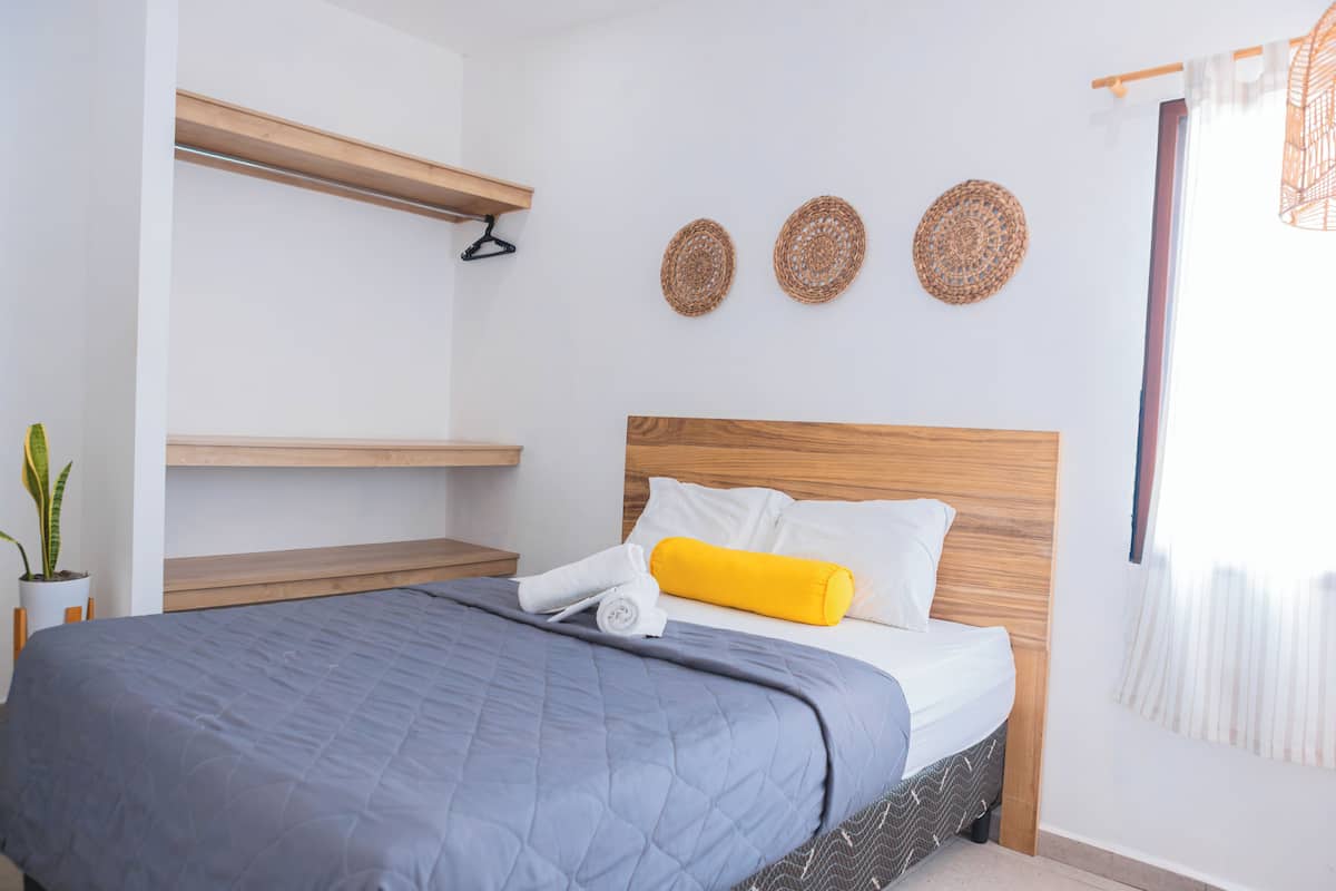 A neatly made bed with a grey quilt, a wooden headboard, and a yellow pillow. There are three woven wall decorations above the bed.