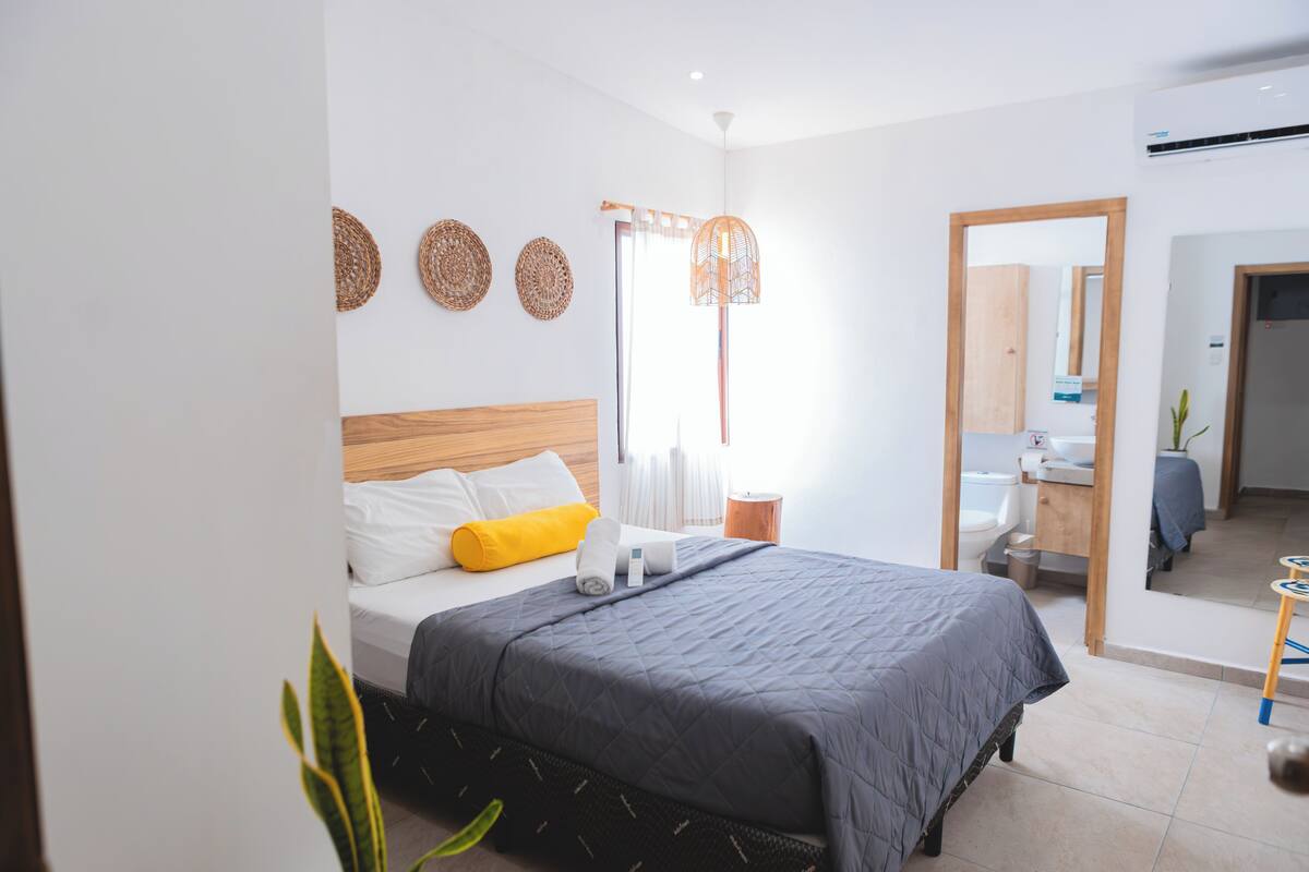 A modern bedroom with a large bed, a bedside table, a mirror, and a bathroom visible through an open door.