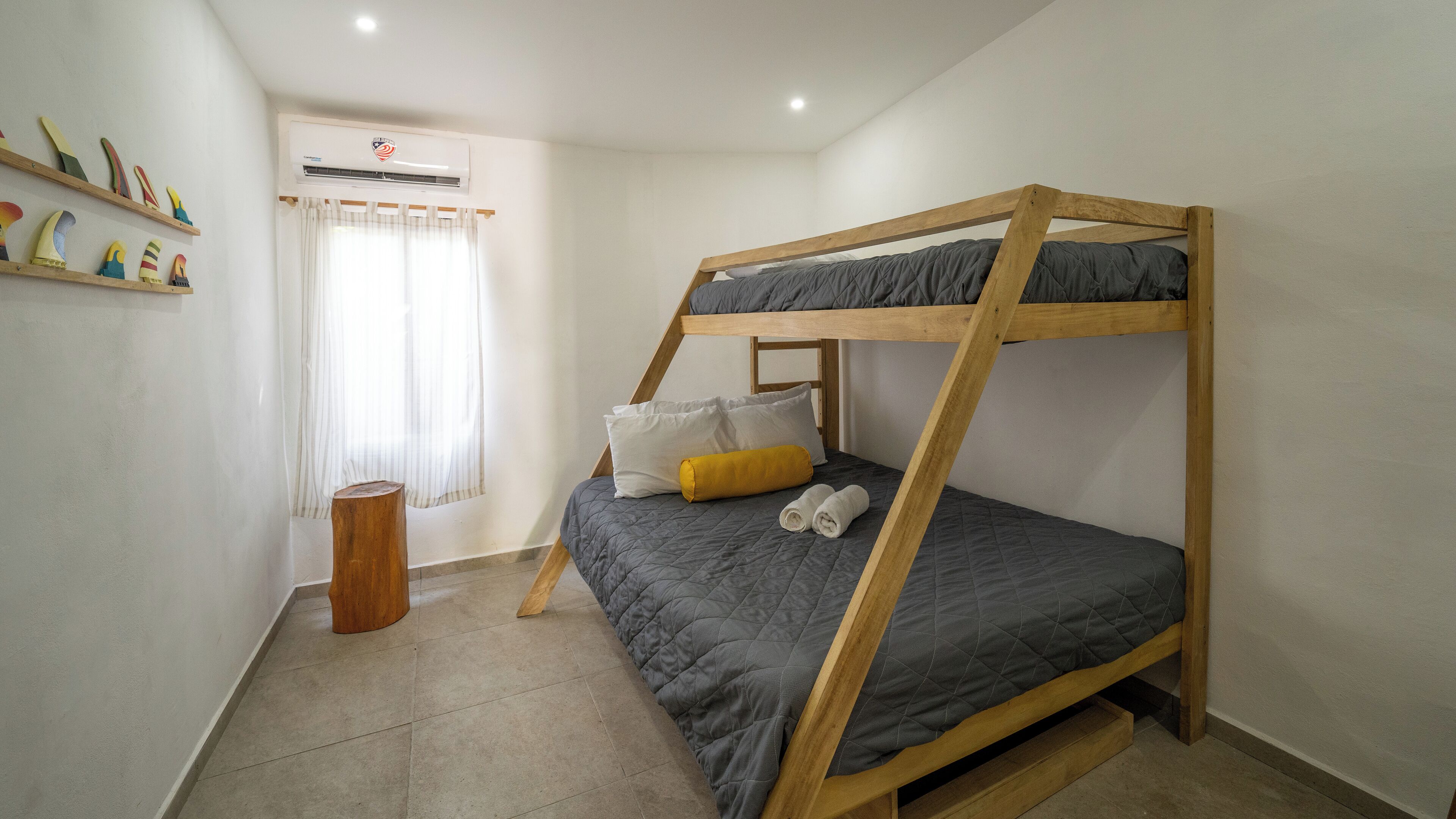Uma cama beliche com um edredom cinza, um banquinho de madeira e uma janela com cortinas.