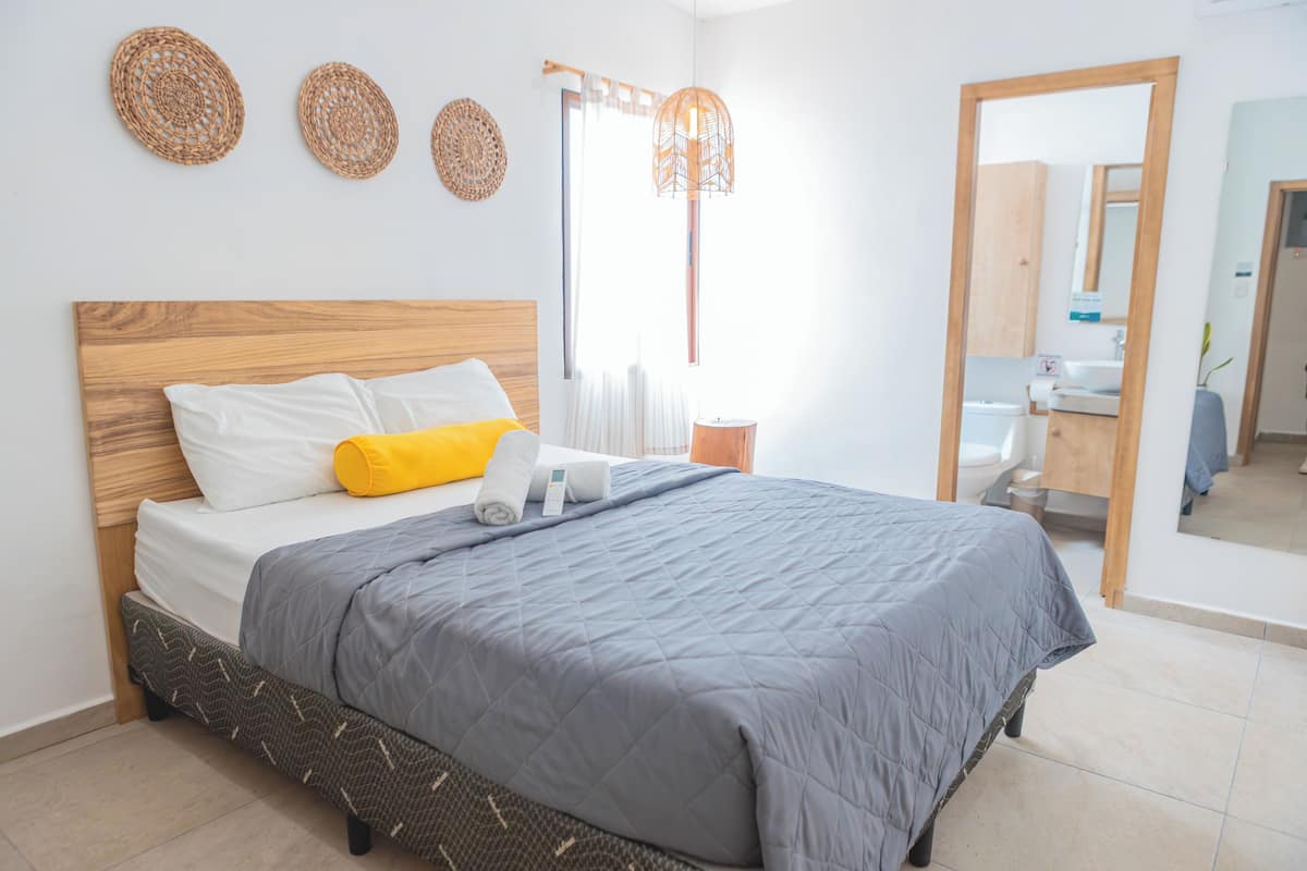 A neatly made bed with a grey quilt, white pillows, and a yellow accent pillow. There are woven wall decorations above the bed and a hanging light fixture.