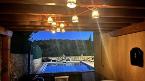 Ein Außenpoolbereich mit Holzbalken und hängenden Lampen, umgeben von Steinmauern und Blick auf Bäume.