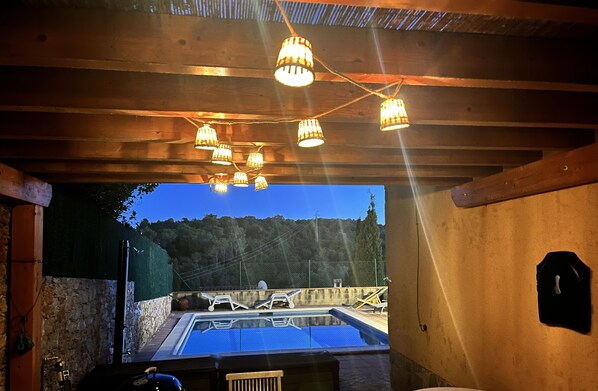 An outdoor swimming pool area with wooden beams and hanging lights, surrounded by stone walls and a view of trees. - Beautiful house with a private pool. BirdsongBegur (Begur)