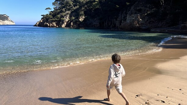 Ein Kind geht am sandigen Strand entlang, im Hintergrund sind steile Felsen und klares, blaues Wasser zu sehen.