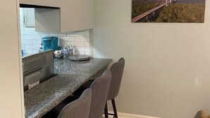 Une cuisine de taille réduite, dotée d’un plan de travail en granit, de quatre chaises grises, d’une machine à café et d’un tableau représentant un pont au-dessus de l’eau, accroché au mur.