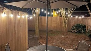Une terrasse extérieure avec une table ronde et des chaises, un parasol et des guirlandes lumineuses.