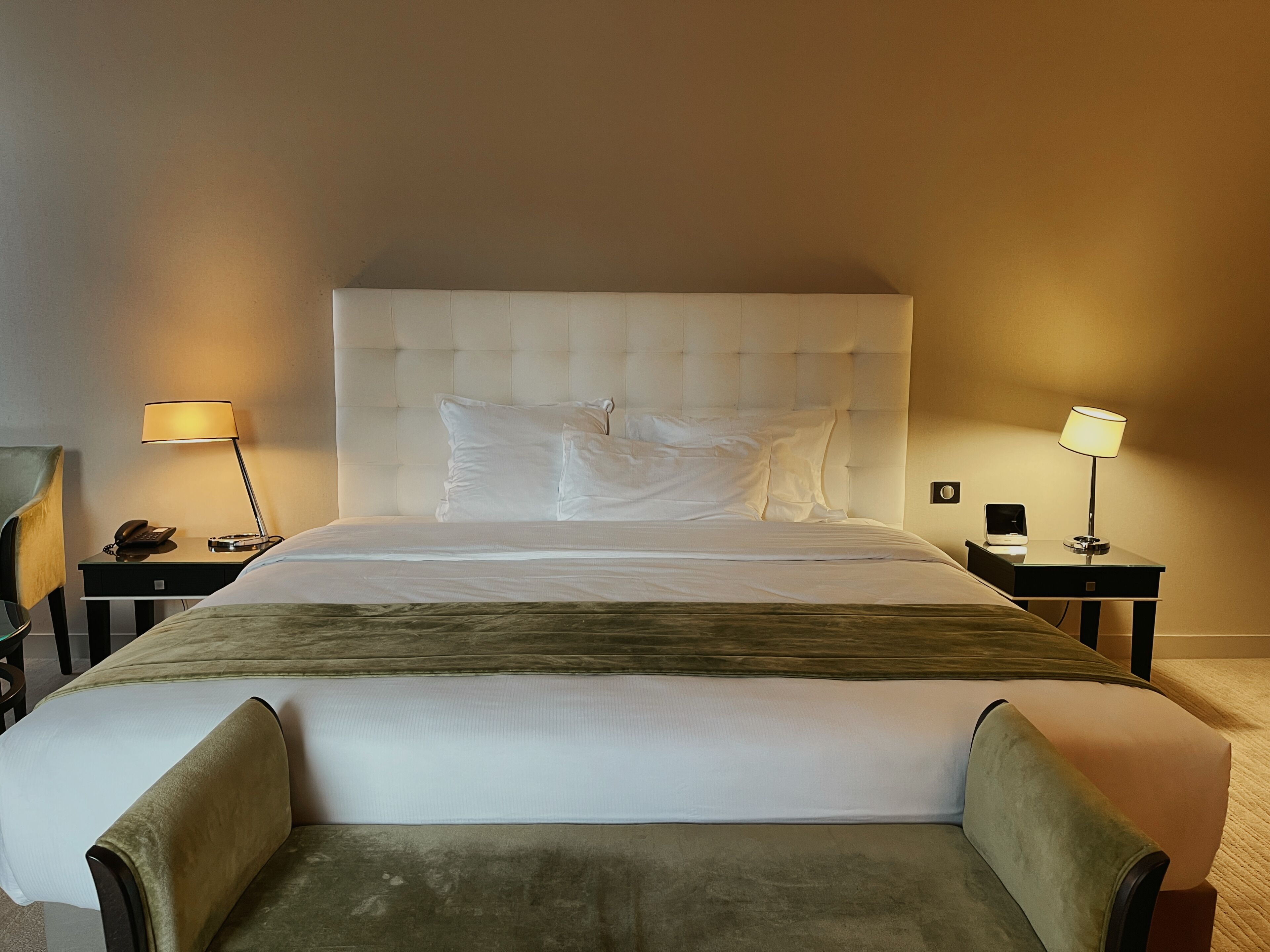Cama bem arrumada com cabeceira estofada, duas mesas de cabeceira com abajures e um banco estofado verde.