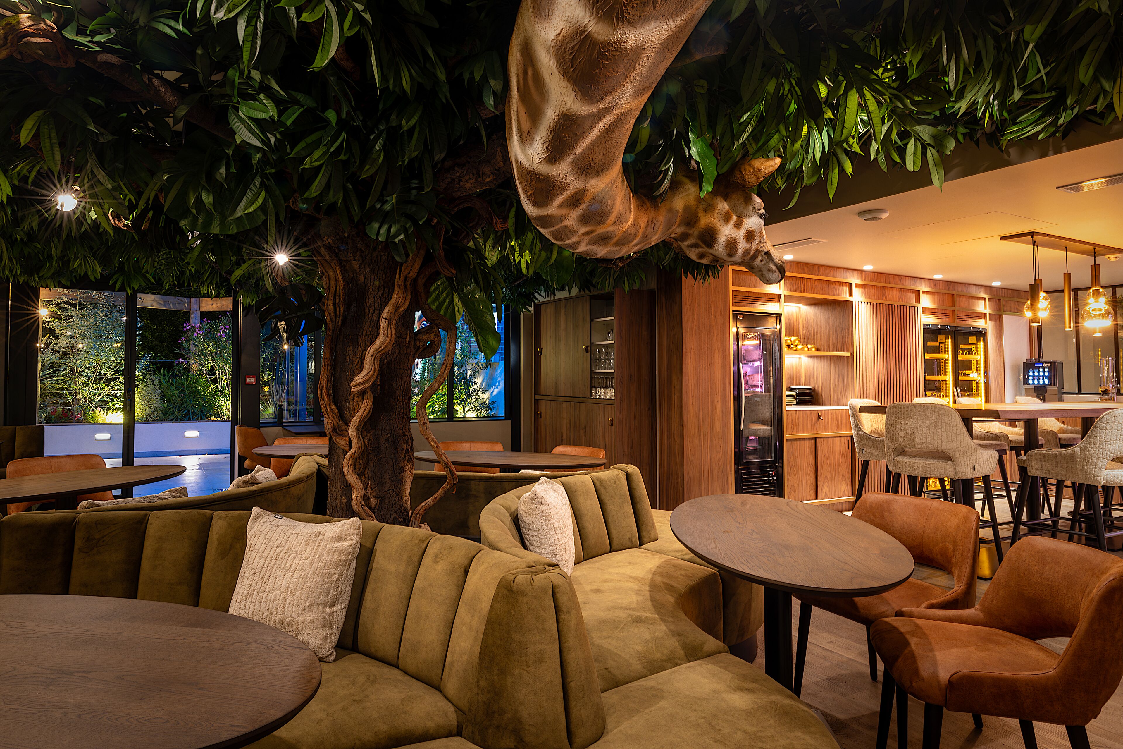 A restaurant with a giraffe sculpture, cushioned seating, and a view of greenery outside.