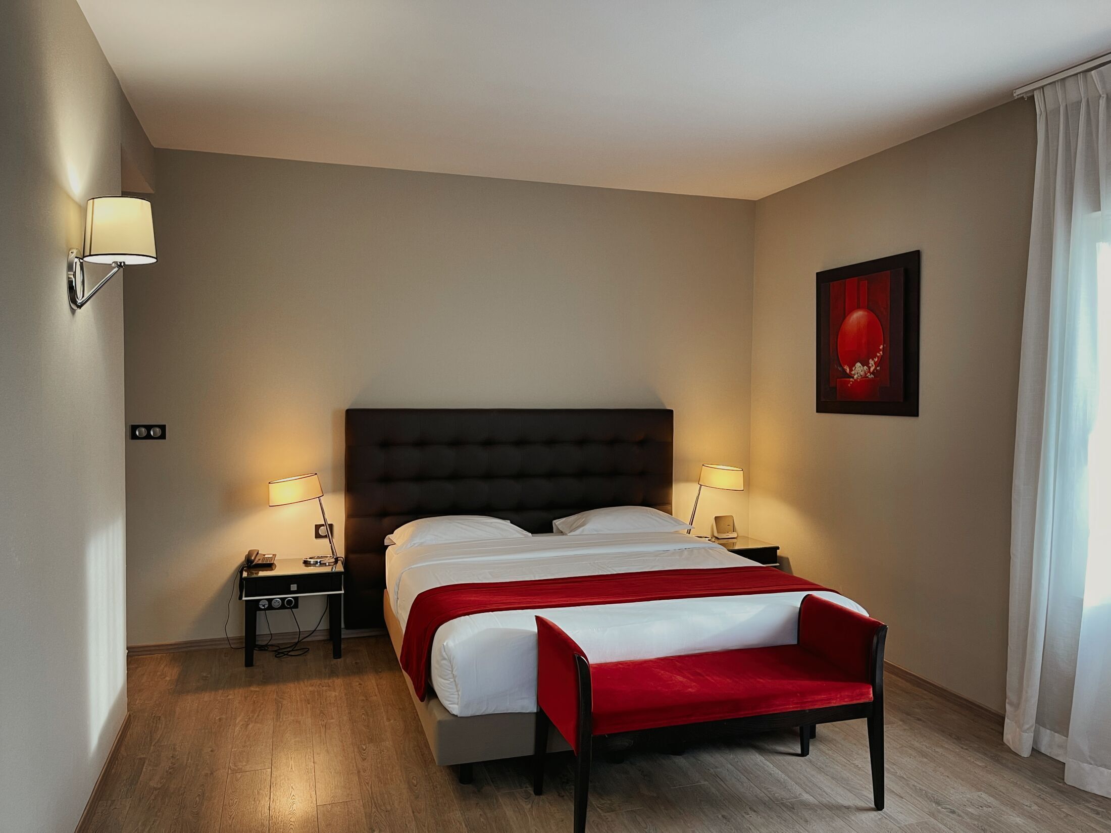 A hotel room with a large bed, two bedside tables, a red bench, a wall-mounted lamp, and a framed picture.