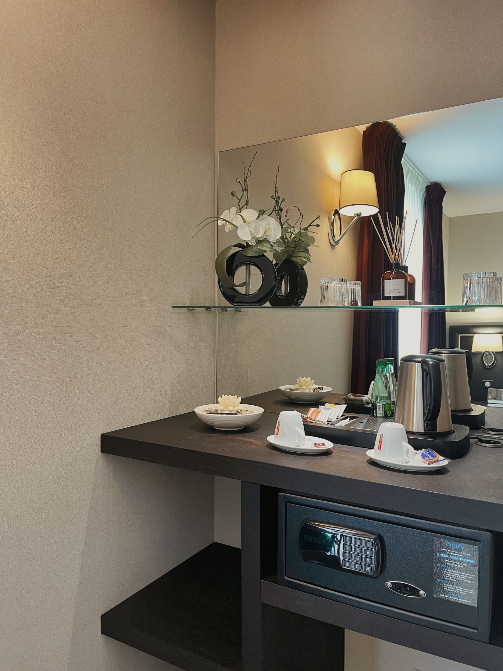 A hotel room with a dark wooden desk, a small shelf with a vase of white flowers, a kettle, a coffee maker, and a digital safe.