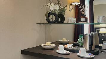 A hotel room with a dark wooden desk, a small shelf with a vase of white flowers, a kettle, a coffee maker, and a digital safe.