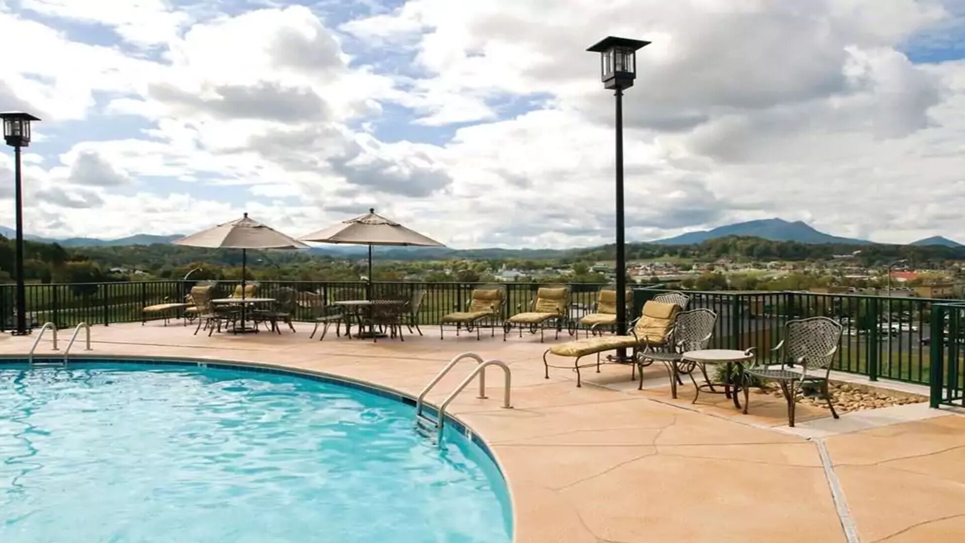 A pool area with lounge chairs and umbrellas, surrounded by a fence with a view of mountains and a town in the background.