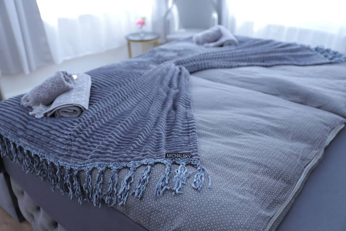 Ein ordentlich bezogenes Bett mit grauer Decke und einem aufgerollten Handtuch auf der Seite.