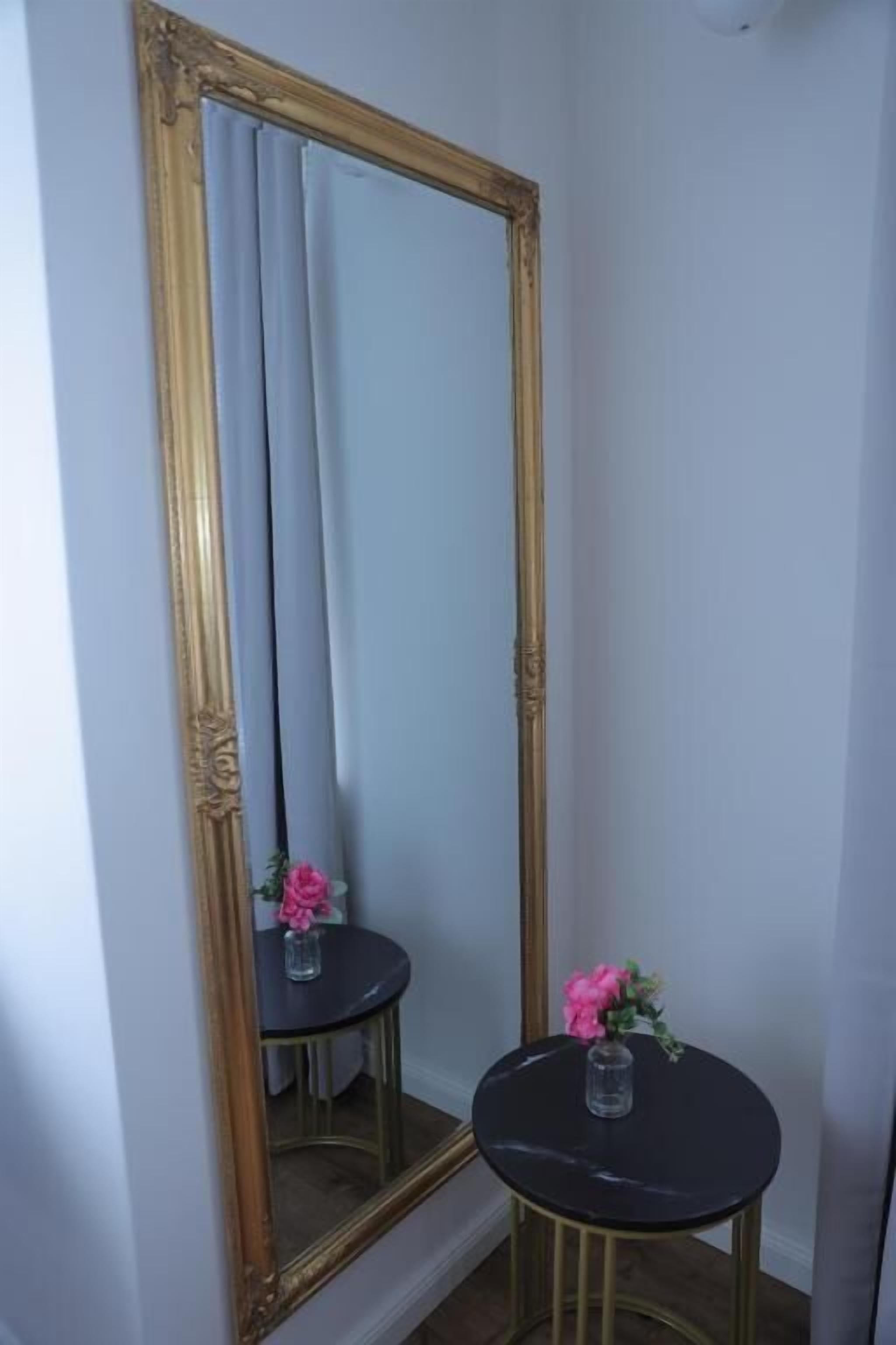 A room with a large mirror, a small round table with a vase of flowers, and a gold-framed mirror.