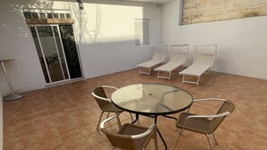 Un patio con una mesa redonda de vidrio y sillas de mimbre, una puerta corredera de cristal y un balcón con barandilla de madera.