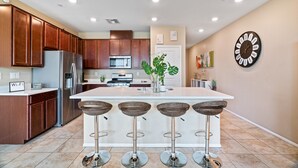 A modern kitchen with wooden cabinets, a stainless steel refrigerator, a large island with bar stools, and a wall clock.