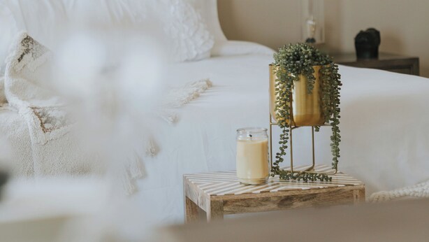A neatly arranged bedroom with a bed, a bedside table with a candle and a vase, a decorative wall panel, and a painting.