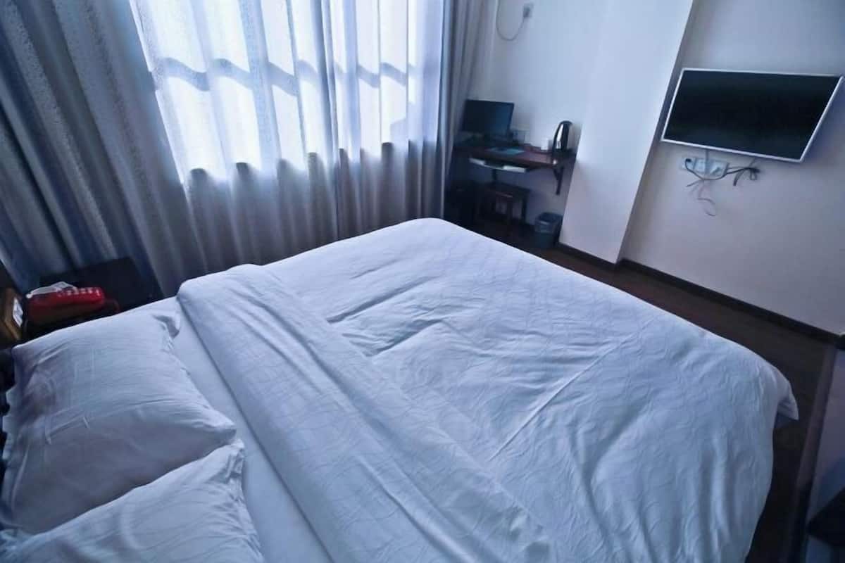 A hotel room with a bed, a desk with a computer, a TV mounted on the wall, and a window with curtains.