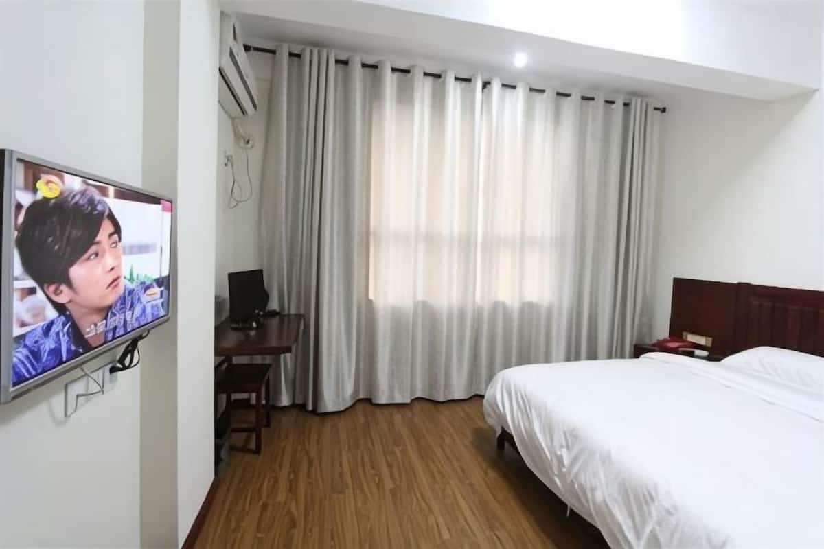 A hotel room with a bed, a desk with a chair, a television mounted on the wall, and a window with curtains.