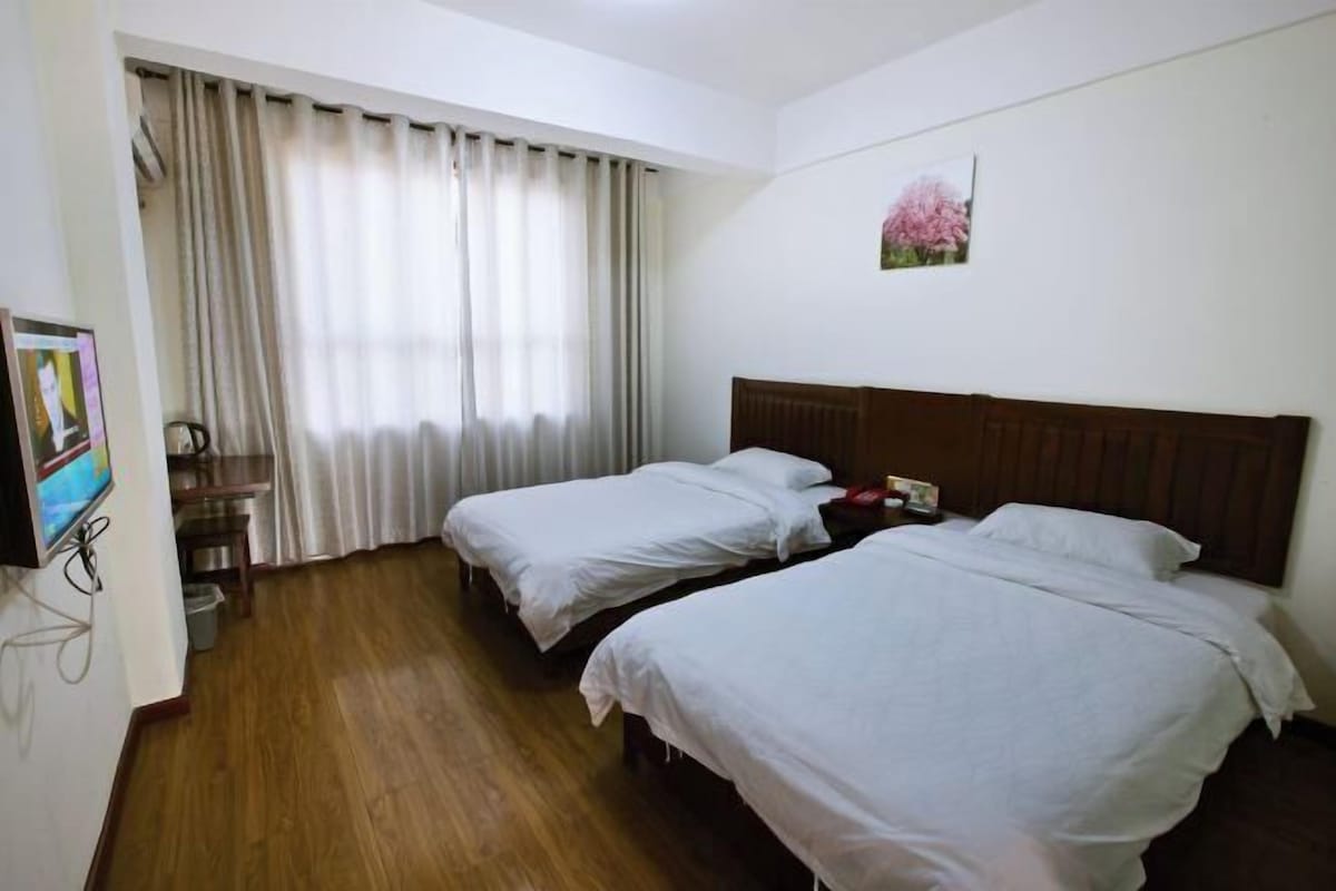Habitación de hotel con dos camas, piso de madera, televisión y ventana con cortinas.
