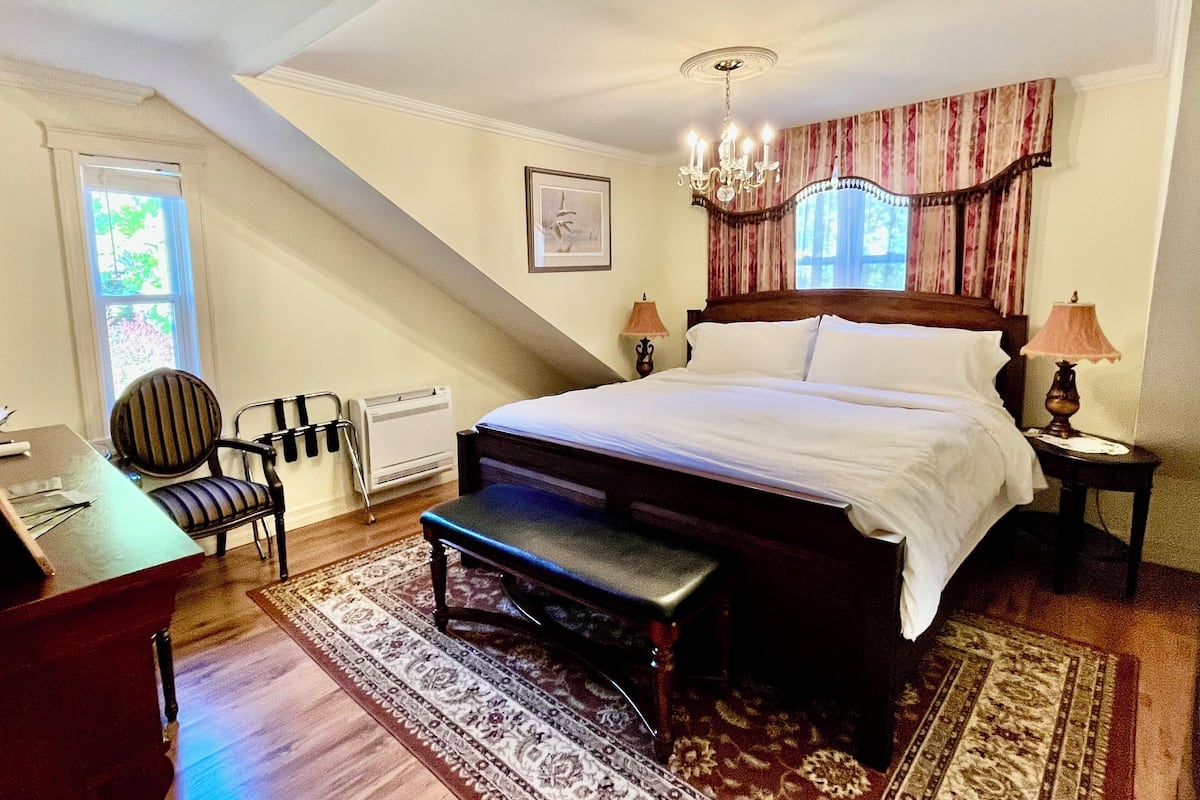 A bedroom with a large bed, a desk, a chair, a bench, a radiator, a chandelier, and a window with curtains.