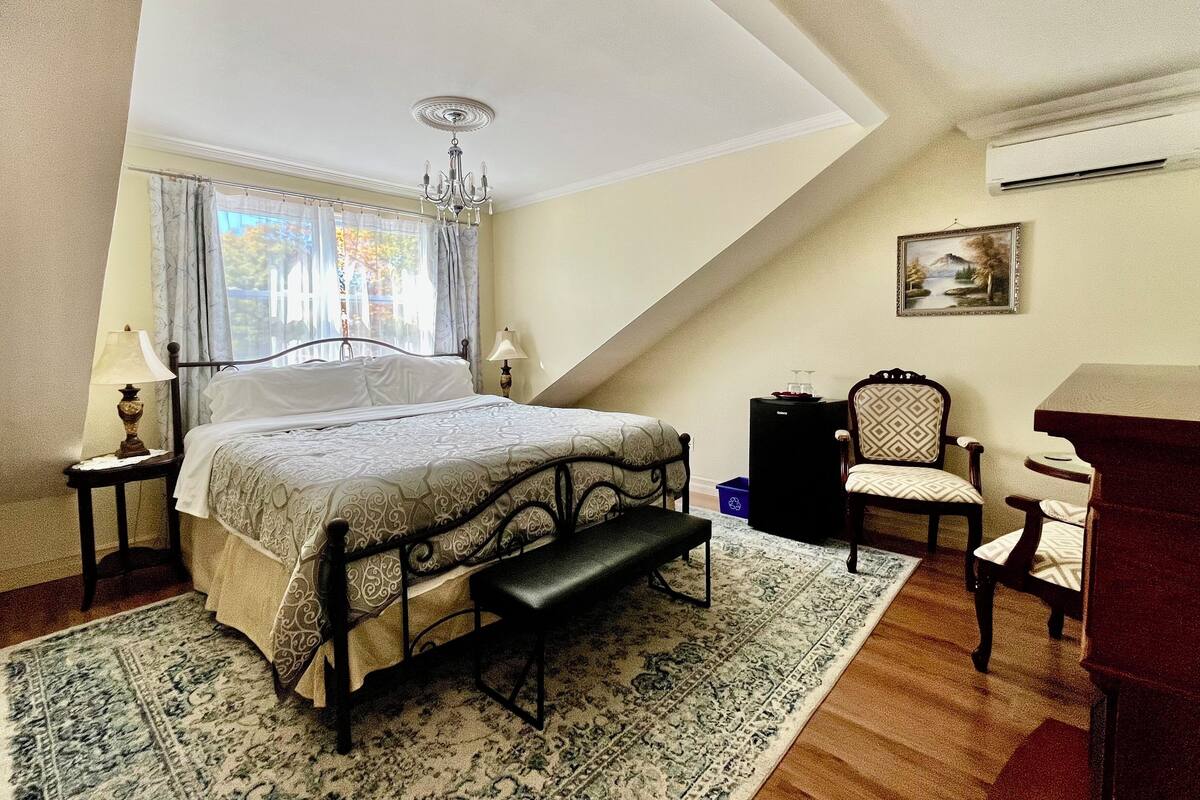A bedroom with a bed, bedside lamps, a bench, a chair, a desk, and a painting on the wall.