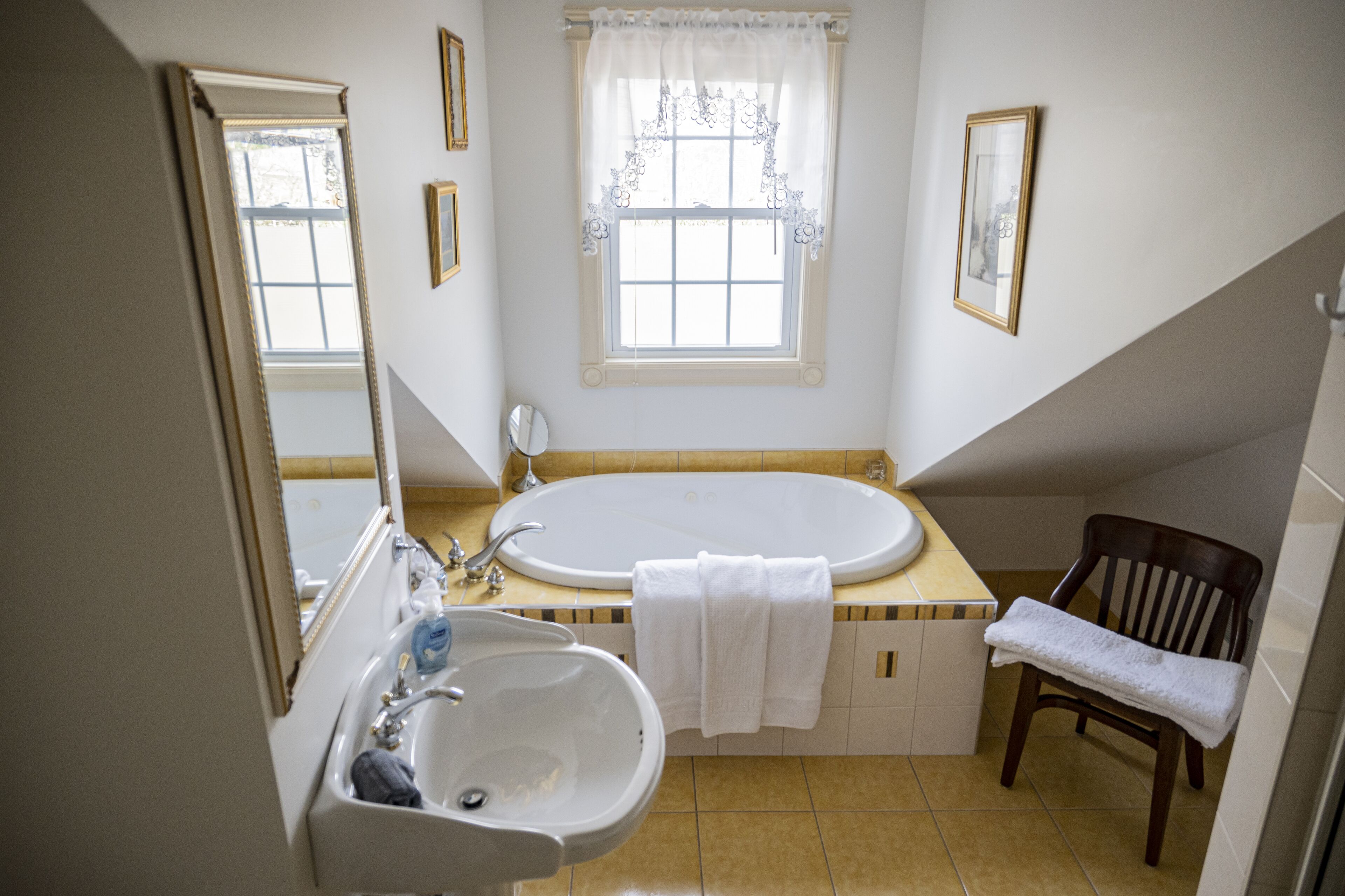 Un baño con bañera, lavabo, espejo, ventana con cortina y una silla con toalla.