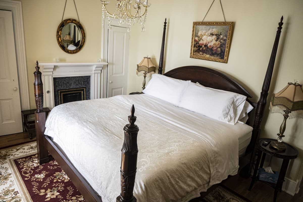 A bedroom with a four-poster bed, a fireplace, a chandelier, and a painting on the wall.
