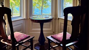 Ein Zimmer mit Erkerfenster und Blick auf ein Gewässer und Bäume im Herbstlaub. Vor dem Fenster steht ein runder Tisch mit zwei Stühlen.