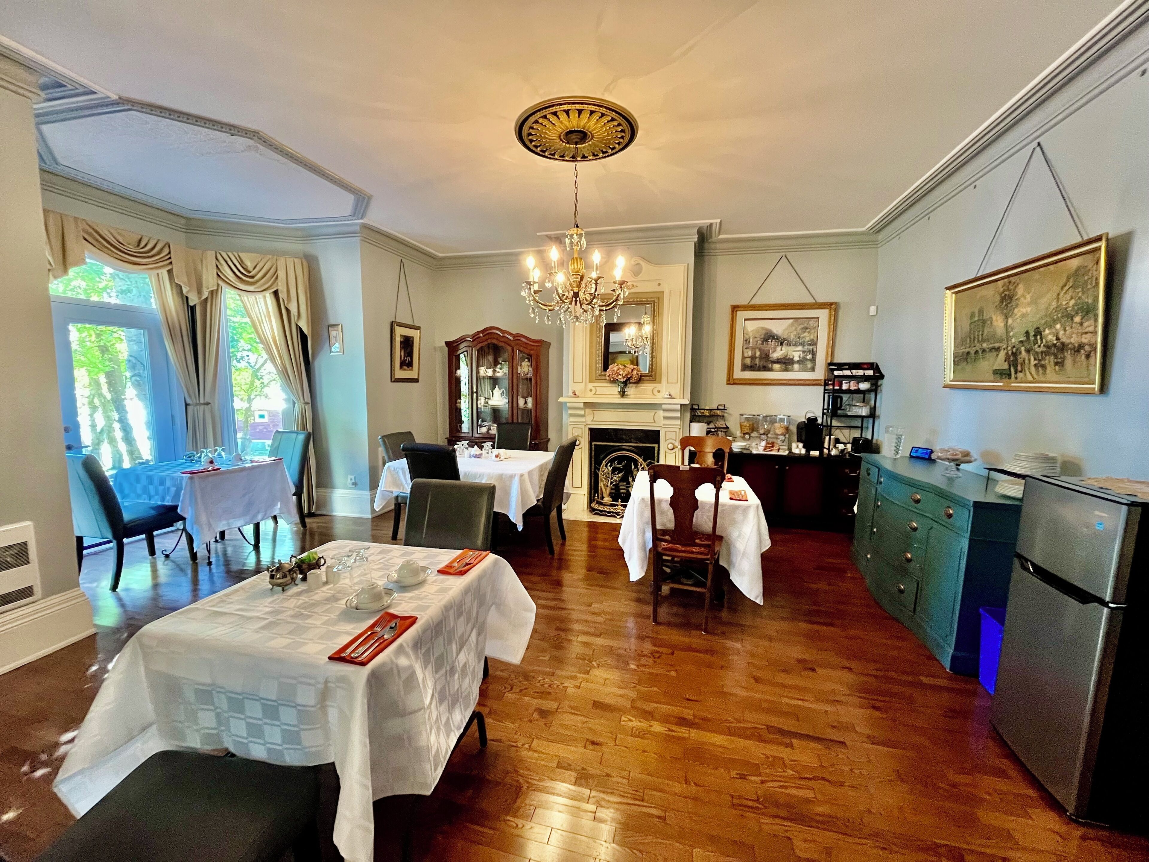 A dining area with a chandelier, wooden dining table, chairs, and a kitchen area with a refrigerator.