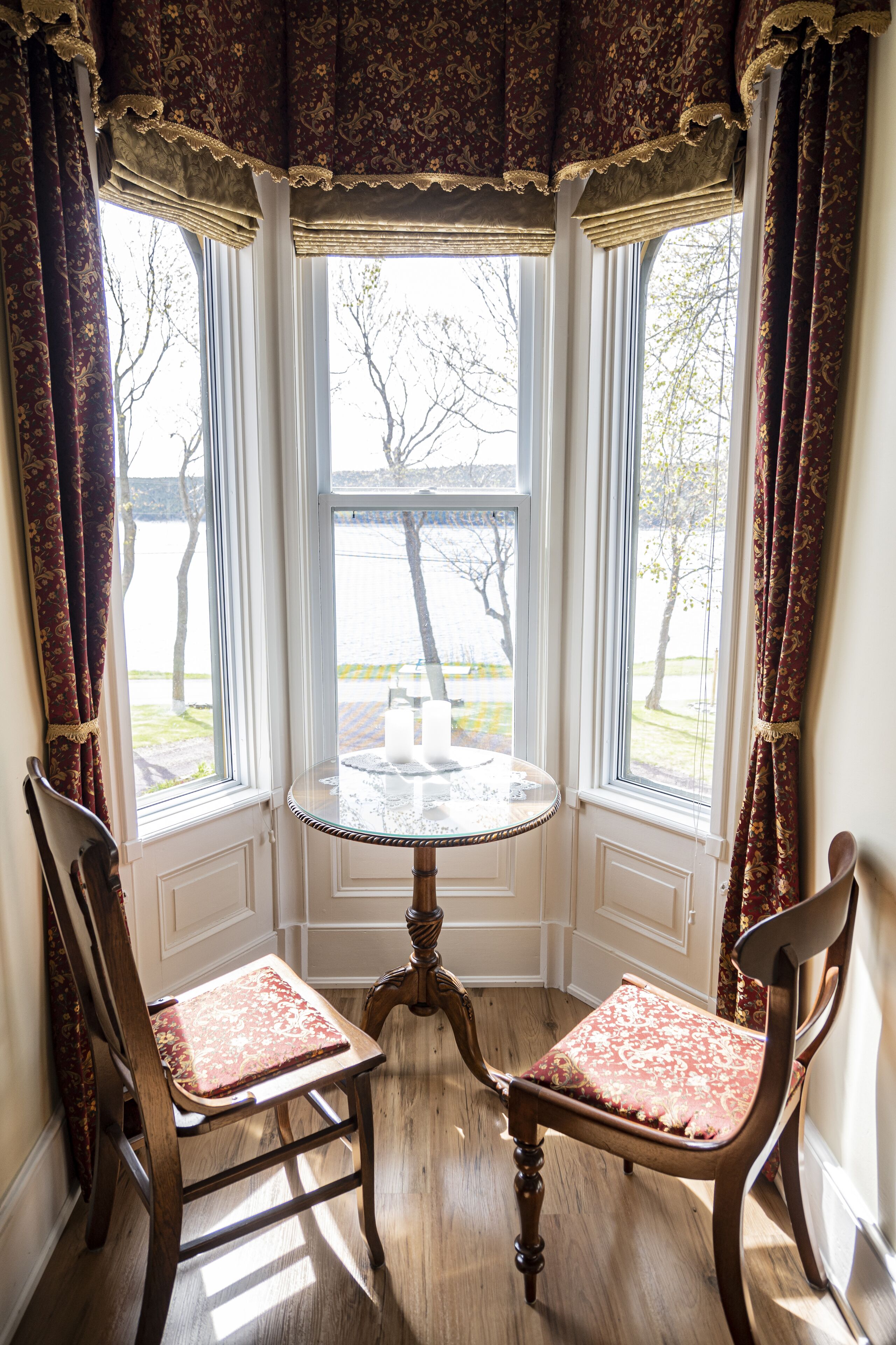 A corner room with two wooden chairs, a round table, and a bay window with curtains.