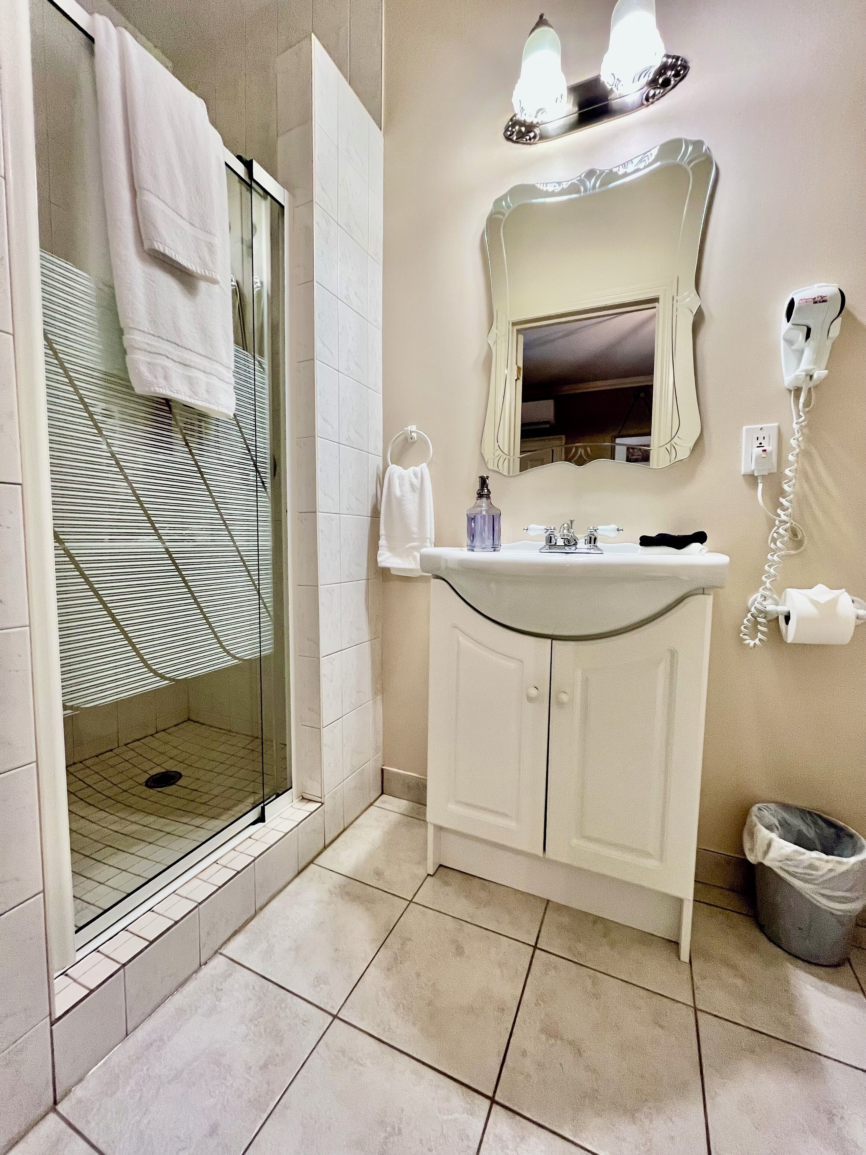 A bathroom with a glass shower enclosure, a white sink with a mirror above it, and a hairdryer mounted on the wall.