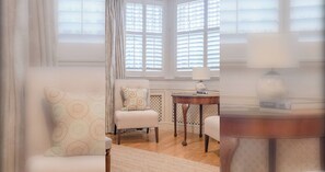 A room with a white armchair, a patterned sofa, a small table with a lamp, and a view through a window with blinds. - Serene Studio Hideaway, Kensington and Chelsea (London)