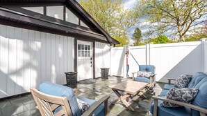 A patio area with a wooden table and blue upholstered chairs, a white fence, and a tree in the background. - Ultra-Modern Studio | Near Hospital & NYCIM | WiFi (Huntington)