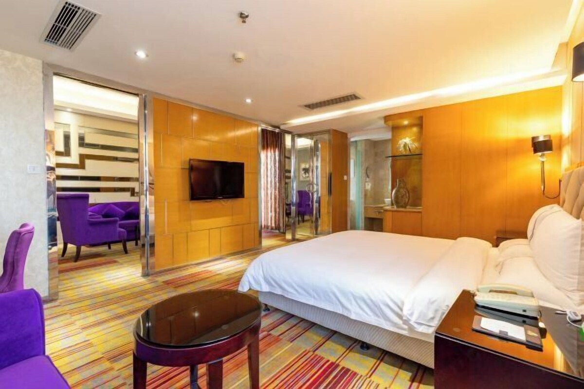 A hotel room with a large bed, a flat-screen TV, a sofa, a chair, and a colorful striped carpet.