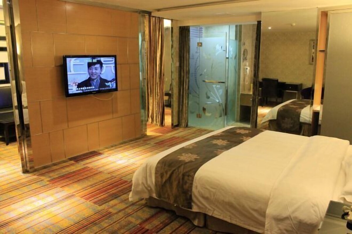 A hotel room with a bed, a television mounted on the wall, and a glass door leading to a bathroom.
