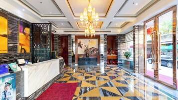 A hotel lobby with a reception desk, a chandelier, a patterned floor, and a large window with a view of the outside.