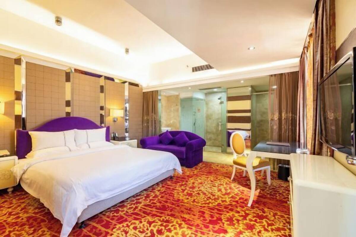 A hotel room with a large bed, a purple armchair, a desk, and a large mirror.