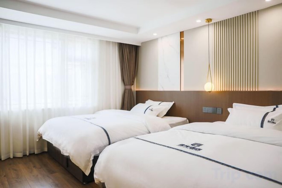 A hotel room with two beds, a large window with curtains, a wall-mounted light, and a wooden headboard.