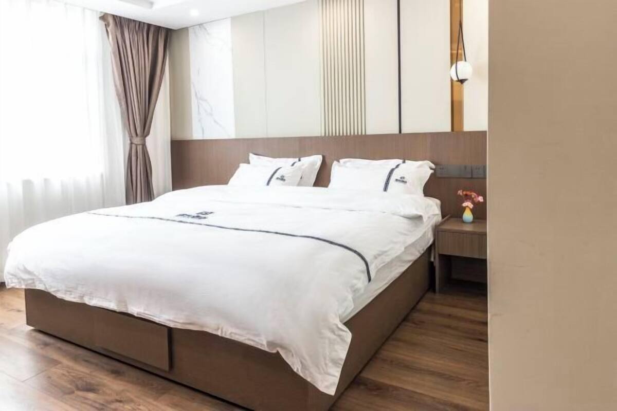 A modern hotel room with a large bed, a nightstand, a mirror, and a window with curtains.