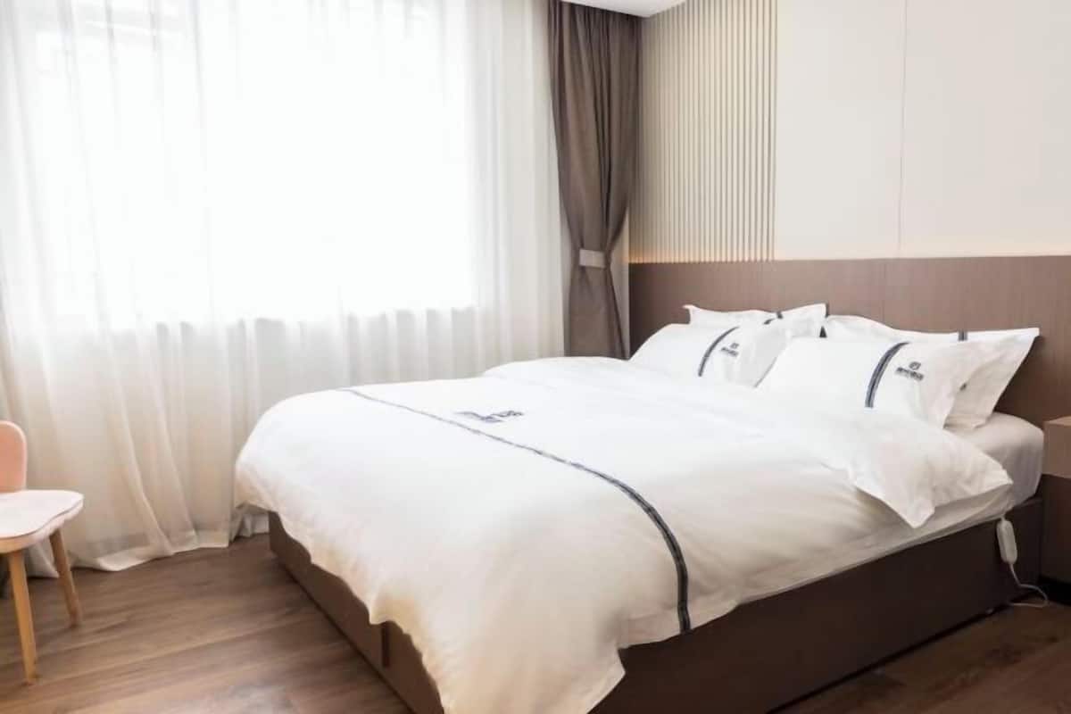 A neatly made bed with white linens and a brown headboard, a wooden chair, and a window with sheer curtains.