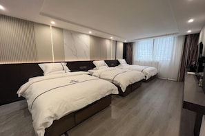 Habitación de hotel con tres camas, cabecero oscuro, ventana grande con cortinas y televisor de pantalla plana.