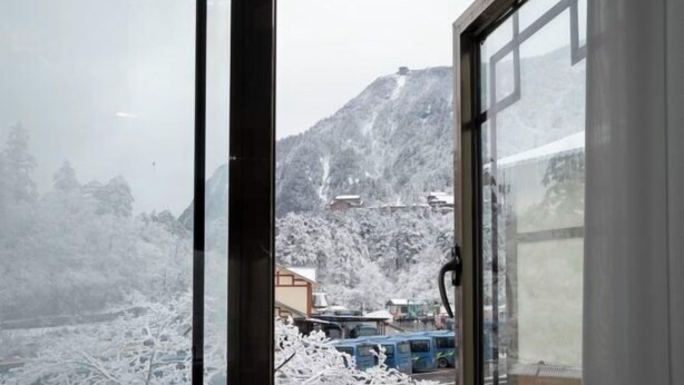 Vista a través de una ventana que muestra una montaña al fondo, un edificio con techo inclinado y un autobús estacionado.
