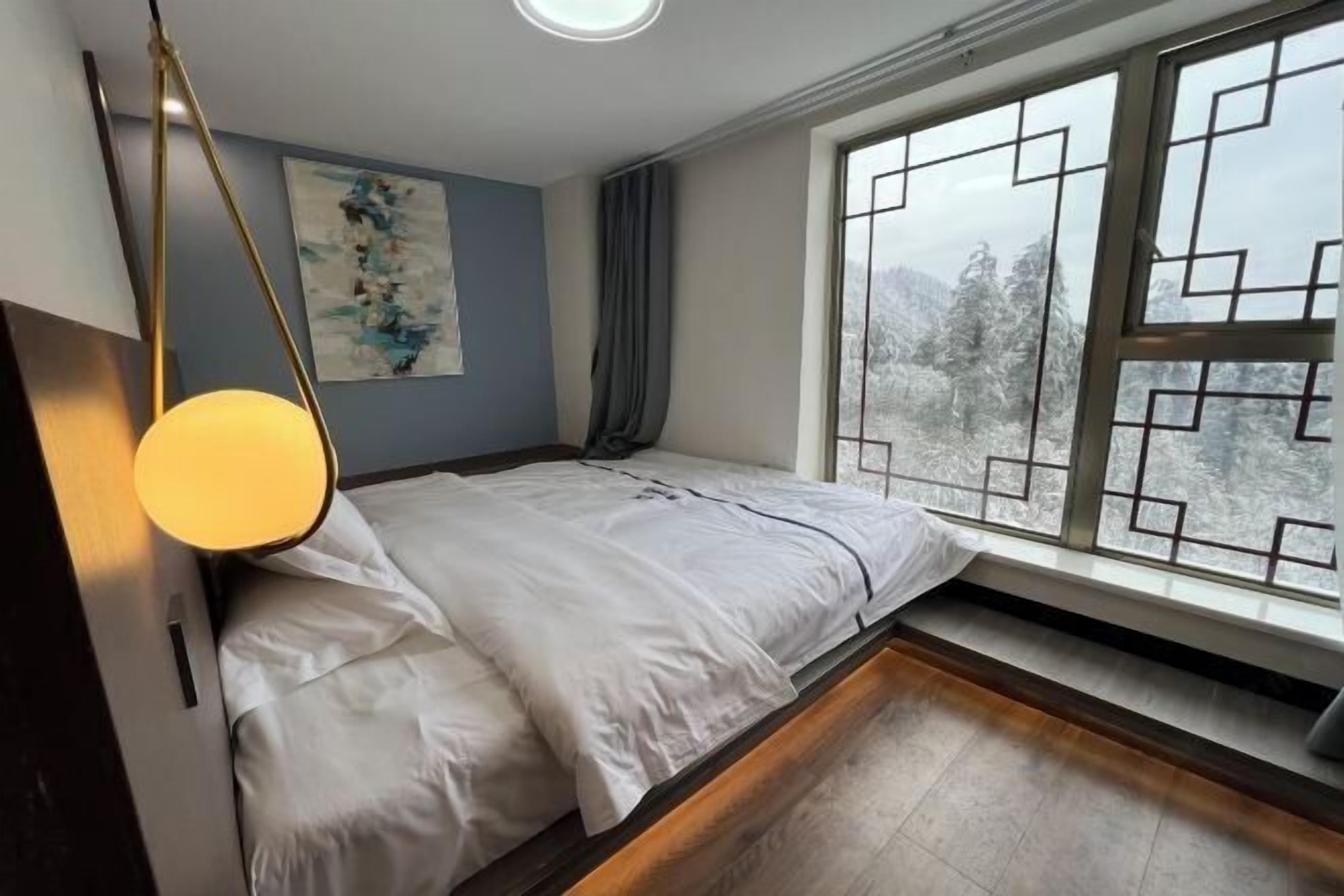 A bedroom with a bed, a window with a view of trees, a painting on the wall, and a lamp.