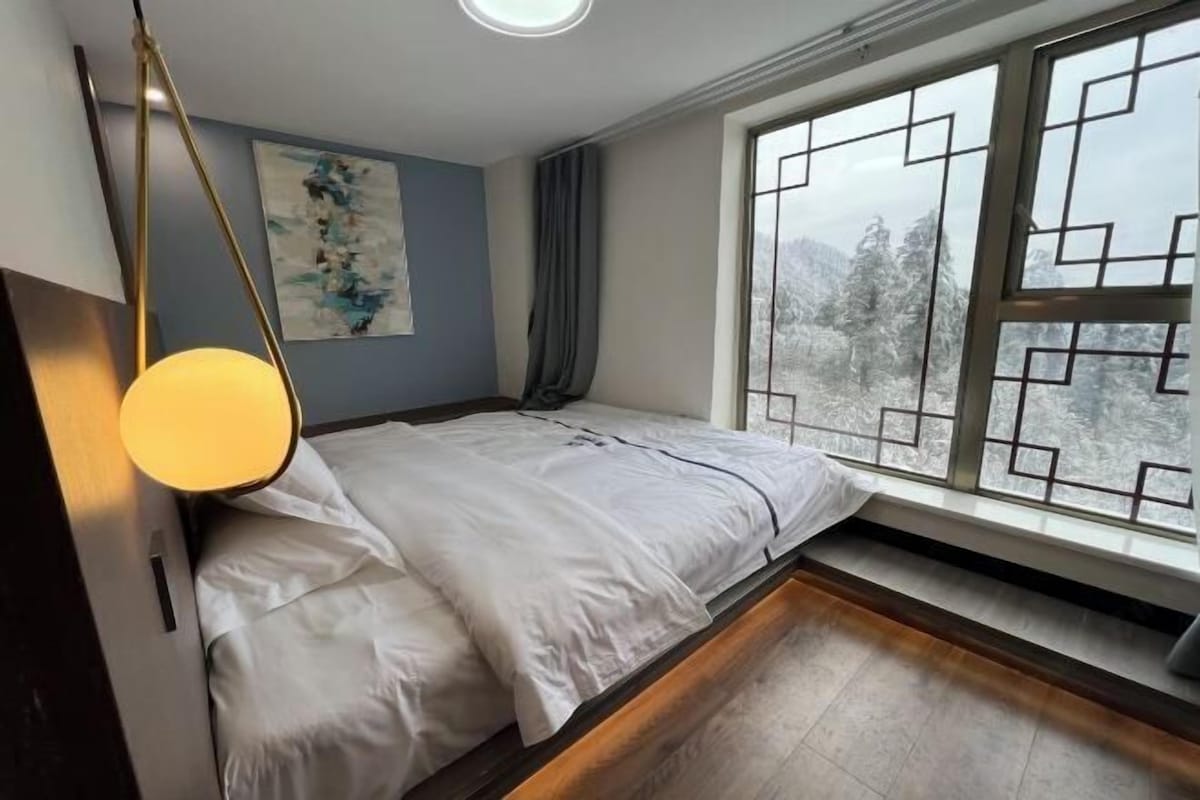 Un dormitorio con cama, ventana con vista a árboles, un cuadro en la pared y una lámpara.